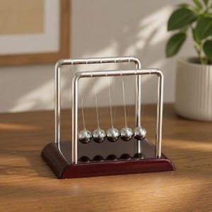 Op de afbeelding: Een Newton's cradle bureau speelgoed met vijf gepolijste stalen ballen die zijn opgehangen aan een verchroomd frame. De basis is van donker, gebeitst hout. Het speelgoed demonstreert de behoud van impuls en energie.