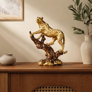 May include: A gold-colored leopard sculpture perched on a textured, brown tree branch. The leopard has a spotted pattern and is posed in a stalking position. The sculpture is a decorative item, suitable for home decor.