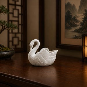 May include: A white ceramic swan figurine with detailed feather textures. The swan has a curved neck and a small, open bowl-like area on its back. The figurine is placed on a dark wooden surface, with a bonsai tree and artwork in the background.