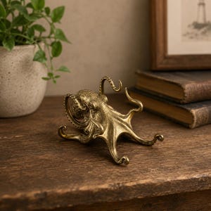 Puede incluir: Una detallada escultura de pulpo de latón con detalles texturizados. La escultura tiene ocho tentáculos y está colocada sobre una superficie de madera. El pulpo es un objeto decorativo, posiblemente un pisapapeles o una pequeña estatua.