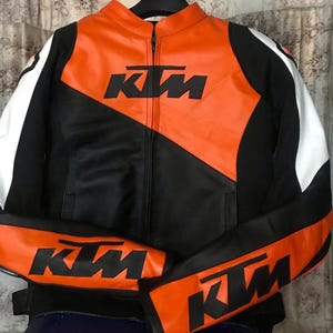 May include: A black, white, and orange motorcycle jacket with the KTM logo. The jacket features a full-length zipper, black sleeves, and orange panels on the chest and arms. The KTM logo is prominently displayed in black.