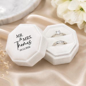 May include: An octagonal white velvet ring box with a lid featuring black printed text that reads MR. & MRS. Thomas 10.12.2026. The open box contains two silver-toned metal rings, one with a single clear stone setting and one plain band.