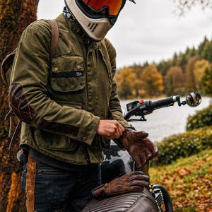 Può includere: Giacca da moto verde oliva con toppe sui gomiti e dettagli in pelle marrone. La giacca ha più tasche e una piccola toppa con logo. La persona indossa guanti in pelle marrone e un casco bianco con visiera arancione. Una moto è sullo sfondo.