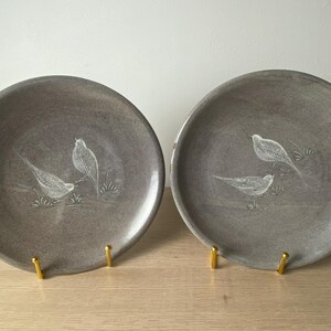 May include: Two round ceramic plates with a smooth, muted gray glaze. Each plate features a white, etched illustration of two small birds perched among blades of grass. The plates are displayed upright on small, gold-toned metal stands.