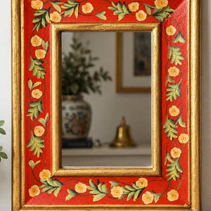 May include: A rectangular mirror featuring a wide, red-painted frame decorated with hand-painted yellow flowers and green leaves. The frame has a gold-toned inner and outer border, providing a metallic finish to the edges of the piece.