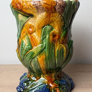 May include: A ceramic vase featuring a raised, three-dimensional relief of yellow daffodils and green foliage. The surface has a visible crackle glaze finish. The base of the vase is a deep blue color, and the overall design is textured and glossy.