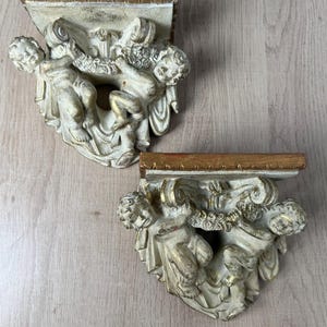 May include: Two wall brackets featuring sculpted cherubs in a cream and gold finish. Each bracket has a flat rectangular top shelf and a decorative base adorned with two figures holding a garland, showcasing intricate carved details and textures.