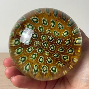Puede incluir: Un pisapapeles de vidrio esf&eacute;rico con una base de color &aacute;mbar miel, incrustado con un patr&oacute;n repetitivo de peque&ntilde;as varillas millefiori florales de varias capas en tonos blanco, verde y azul oscuro, creando un dise&ntilde;o de mosaico denso.