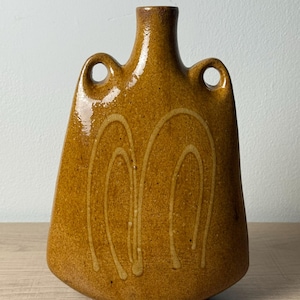 May include: A mustard-colored ceramic vase with a glossy finish, featuring a narrow neck, two integrated side handles, and a flat base. The body is decorated with two light-colored, hand-painted arch designs on the front surface.