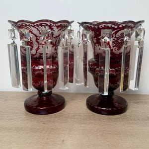 May include: Two ruby red glass vases with crystal accents. The vases have a scalloped rim and etched floral designs. Rectangular crystal prisms hang from the rim. The vases are on a wooden surface.