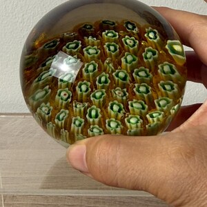 Puede incluir: Un pisapapeles de vidrio esf&eacute;rico con una capa exterior transparente y una disposici&oacute;n interna de numerosos peque&ntilde;os patrones florales millefiori repetidos en tonos verde, blanco y amarillo sobre un fondo marr&oacute;n dorado.