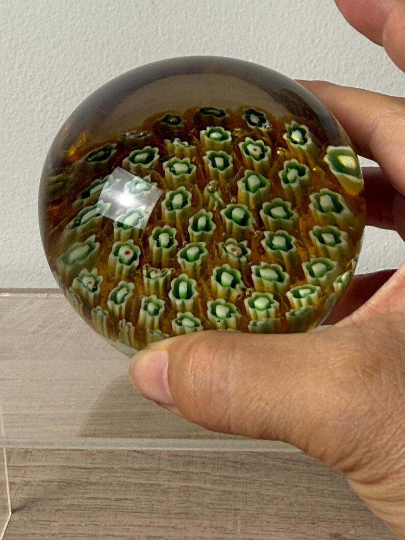 Puede incluir: Un pisapapeles de vidrio esf&eacute;rico con una capa exterior transparente y una disposici&oacute;n interna de numerosos peque&ntilde;os patrones florales millefiori repetidos en tonos verde, blanco y amarillo sobre un fondo marr&oacute;n dorado.