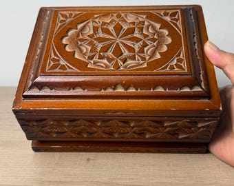 Puede incluir: Una caja rectangular de madera con acabado marrón medio, que presenta intrincadas tallas geométricas en la tapa y los laterales. La tapa muestra un patrón central en forma de estrella rodeado de bordes festoneados y un motivo triangular.