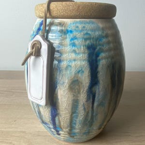 May include: A ceramic jar with a crackle glaze finish in shades of cream, blue, and brown. The jar features a cylindrical cork lid and a small, rectangular white ceramic tag attached to the neck with a thin leather cord, creating a textured appearance.