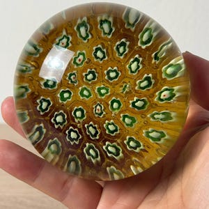 Puede incluir: Un pisapapeles de vidrio redondo con una base de color &aacute;mbar miel y un patr&oacute;n de panal incrustado de peque&ntilde;as varillas de vidrio millefiori en forma de flor blanca y verde dispuestas en una cuadr&iacute;cula geom&eacute;trica repetitiva.