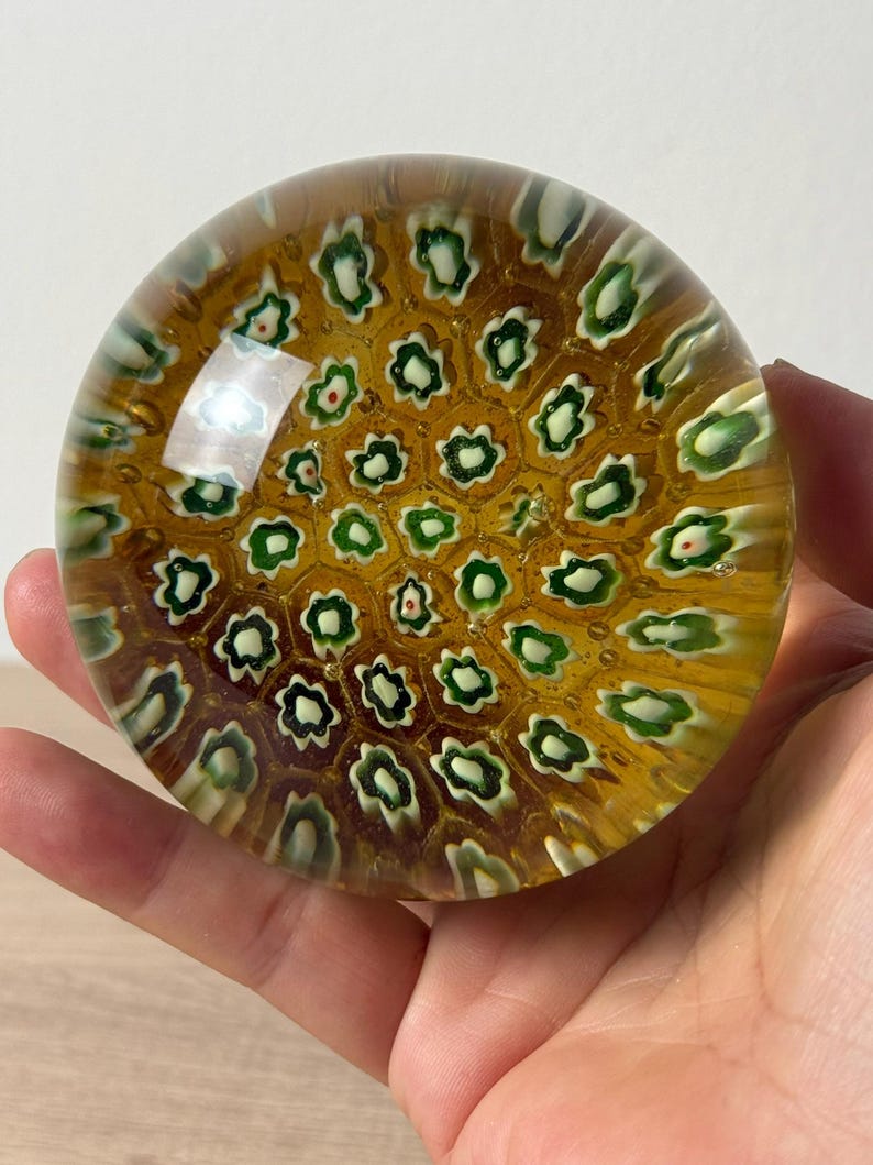 Puede incluir: Un pisapapeles de vidrio redondo con una base de color &aacute;mbar miel y un patr&oacute;n de panal incrustado de peque&ntilde;as varillas de vidrio millefiori en forma de flor blanca y verde dispuestas en una cuadr&iacute;cula geom&eacute;trica repetitiva.