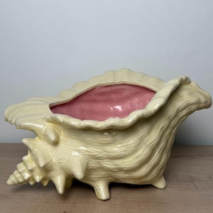 May include: A large, ceramic seashell planter in a light yellow color with a pink interior. The planter has a textured surface and is supported by three small feet. The shell design features intricate details and a ruffled edge.