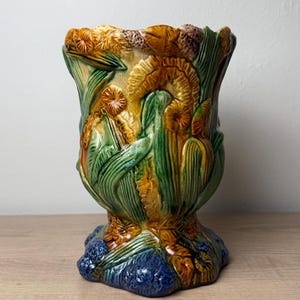 May include: A ceramic vase featuring a high-relief, three-dimensional design of yellow and orange flowers with green leaves. The base of the vase is textured and colored in a deep blue, while the upper body displays a glossy, multi-colored glaze.
