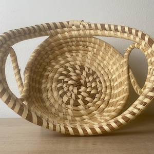 May include: A round woven basket featuring a coiled design with alternating light beige and dark brown strips. The basket has two large, arched handles integrated into the rim, creating a symmetrical shape with a deep, circular interior basin.
