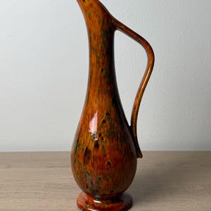 May include: A tall ceramic pitcher with a slender neck and a single handle. The surface features a glossy, mottled glaze in shades of deep amber and brown, accented with small, scattered flecks of bright green throughout the entire body.