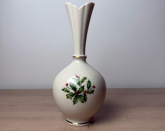 May include: A cream-coloured ceramic bud vase with a fluted rim and a rounded body. The vase features a painted holly leaf and red berry design on the front, accented by thin gold bands around the neck and the base of the vessel.
