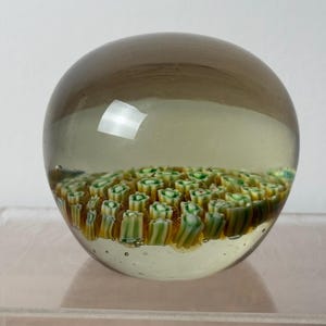 Puede incluir: Un pisapapeles de vidrio esf&eacute;rico con una capa exterior transparente te&ntilde;ida de marr&oacute;n claro. Dentro de la esfera, una densa disposici&oacute;n de peque&ntilde;as varillas de vidrio millefiori cil&iacute;ndricas en tonos blanco, verde y amarillo.