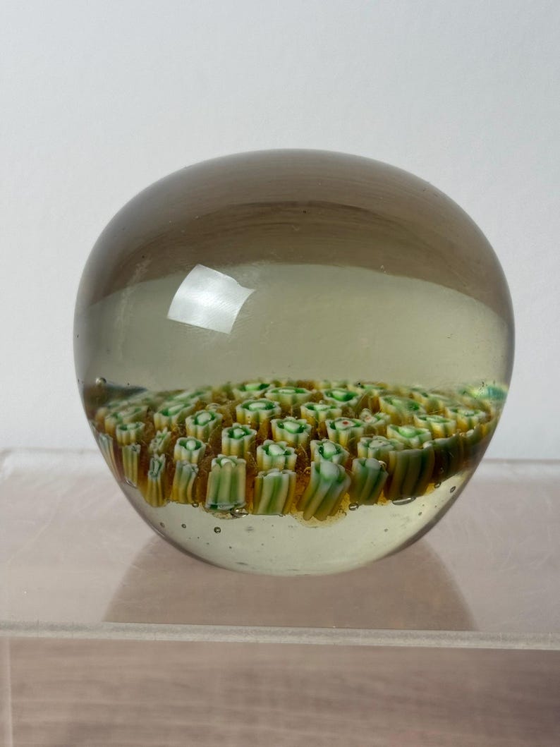 Puede incluir: Un pisapapeles de vidrio esf&eacute;rico con una capa exterior transparente te&ntilde;ida de marr&oacute;n claro. Dentro de la esfera, una densa disposici&oacute;n de peque&ntilde;as varillas de vidrio millefiori cil&iacute;ndricas en tonos blanco, verde y amarillo.
