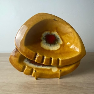 May include: A triangular ceramic ashtray featuring a glossy mustard yellow glaze with a central burst of deep red and dark green. The piece has molded indentations along the rim for resting items and is displayed on a small gold metal stand.