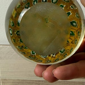 Puede incluir: Un pisapapeles de vidrio transparente con forma esf&eacute;rica que presenta un patr&oacute;n millefiori interno. El dise&ntilde;o consta de numerosos motivos peque&ntilde;os y repetitivos similares a flores en tonos verde y blanco sobre un fondo amarillo dorado.