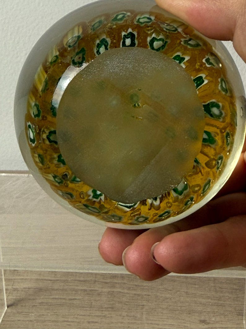 Puede incluir: Un pisapapeles de vidrio transparente con forma esf&eacute;rica que presenta un patr&oacute;n millefiori interno. El dise&ntilde;o consta de numerosos motivos peque&ntilde;os y repetitivos similares a flores en tonos verde y blanco sobre un fondo amarillo dorado.