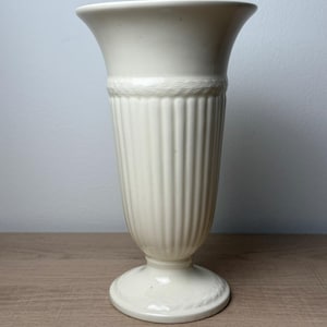 May include: A cream-colored ceramic vase with a flared rim, a vertical ribbed body, and a circular pedestal base. The neck and the base feature a subtle, embossed rope-like texture detail that encircles the circumference of the vessel.