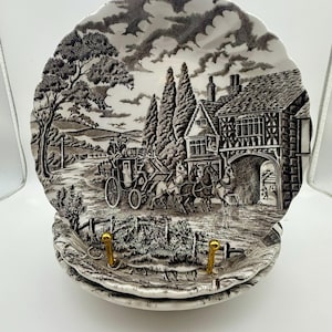 May include: Set of decorative plates with a black and white transferware design. The plates feature a rural landscape scene with a horse-drawn carriage, trees, and a building. The plates have a scalloped edge and gold-colored hooks.