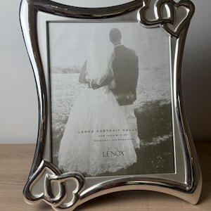 May include: A silver-toned metal picture frame with a curved, wavy border and two interlocking heart accents at the top right and bottom left corners. The frame contains a display insert with the text LENOX PORTRAIT GALLERY FOR YOUR 8 X 10 PERSONAL EXPRESSIONS LENOX Gifts That Celebrate Life.