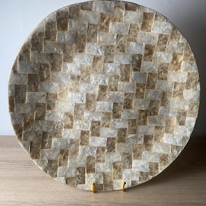 May include: A circular decorative plate featuring a mosaic pattern of rectangular tiles in alternating shades of iridescent white and translucent golden brown. The surface is composed of small, smooth, overlapping pieces arranged in a grid.
