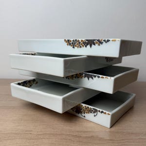 May include: A stack of four rectangular white ceramic trays with low walls. Each tray features a decorative pattern of small brown, gold, and dark gray leaves and flowers along one corner edge, creating a repeating motif across the stacked set.