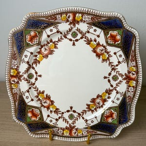 May include: A square ceramic plate with scalloped edges featuring a central white field decorated with a brown floral garland of roses and yellow blossoms. The corners are accented with deep blue, green, and orange floral patterns with gold trim.