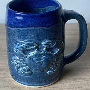 May include: A cylindrical ceramic mug featuring a deep blue glaze with a lighter blue, textured finish. The front of the mug displays a raised, embossed crab design. The mug has a sturdy handle on the right side and a plain, unglazed white base.