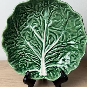 May include: A green ceramic plate shaped like a cabbage leaf with a scalloped edge and a prominent white central vein. The surface has a glossy finish with textured, raised details mimicking the natural appearance of a leafy vegetable.