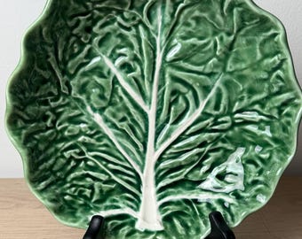 Puede incluir: Un plato de cerámica verde con forma de hoja de col, con un borde festoneado y una nervadura central blanca prominente. La superficie tiene un acabado brillante con detalles texturizados en relieve que imitan la apariencia natural.
