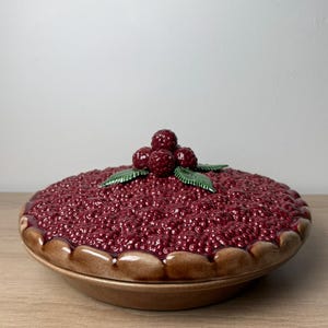 May include: A round ceramic pie dish with a lid featuring a textured, deep red raspberry filling design. The lid is topped with a cluster of sculpted raspberries and two green leaves. The base of the dish has a smooth, light brown crust edge.
