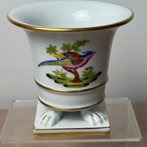 May include: White ceramic vase with a colorful bird illustration and gold trim. The vase features a flared top, a cylindrical body, and a square base with claw feet. The bird has blue, yellow, pink, and green feathers. The vase is approximately 6 inches tall.