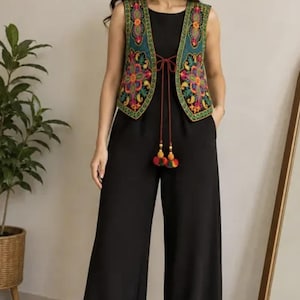 May include: A black sleeveless jumpsuit featuring wide-leg trousers and a round neckline. It is paired with a teal vest adorned with vibrant, multi-colored floral embroidery, a thin tie closure at the center, and decorative hanging tassels.