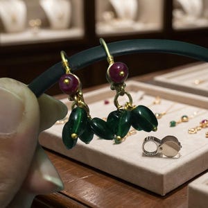 Puede incluir: Pendientes dorados con un grupo de cuentas ovaladas de color verde oscuro y una cuenta roja redonda. Los pendientes se exhiben sobre una superficie clara, mostrando joyas.