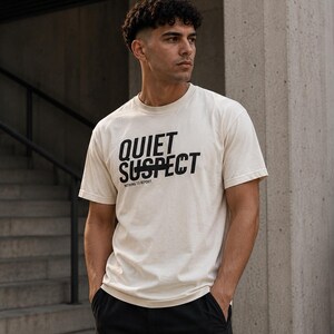 Op de afbeelding: Een crèmekleurig T-shirt met korte mouwen en de zwarte tekst "QUIET SUSPECT" op de borst, met de kleinere tekst "NOTHING TO REPORT" direct onder de hoofdafbeelding in een strak, schreefloos lettertype.
