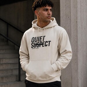 Op de afbeelding: Een beige hoodie met een kangoeroezak aan de voorzijde en lange mouwen. Op de borst staat de zwarte tekst "QUIET SUSPECT" met een horizontale lijn door het woord "SUSPECT" en daaronder de kleinere tekst "NOTHING TO REPORT".