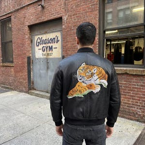 May include: A black leather bomber jacket featuring a large, detailed embroidered patch of a tiger on the back. The jacket has a ribbed black collar and cuffs, and is worn over dark denim jeans, showcasing a classic design with a bold graphic.