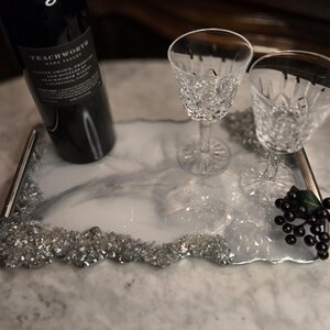 May include: A decorative serving tray with a white and gray marbled design, silver trim, and a sparkling crystal border. The tray holds a bottle of wine and two crystal wine glasses. A cluster of black grapes adds a touch of elegance.