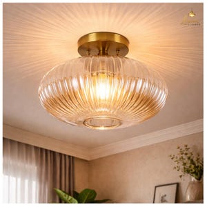 May include: A clear glass ceiling light fixture with a ribbed, rounded shade and a brass-colored base. The light is on, illuminating the room with a warm glow. The fixture is mounted on a white ceiling.