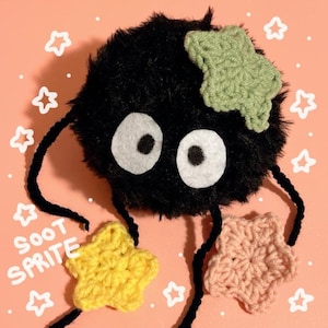 May include: A black, fluffy Soot Sprite plush toy with large white eyes and a green star-shaped crochet detail. The Soot Sprite holds three star-shaped crochet pieces in yellow and pink. The text "SOOT SPRITE" is visible.