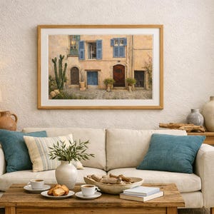 May include: A framed painting of a European street scene hangs above a cream-colored sofa with teal and striped pillows. A wooden coffee table holds a vase, books, and snacks. A lamp and decorative vases sit on side tables.